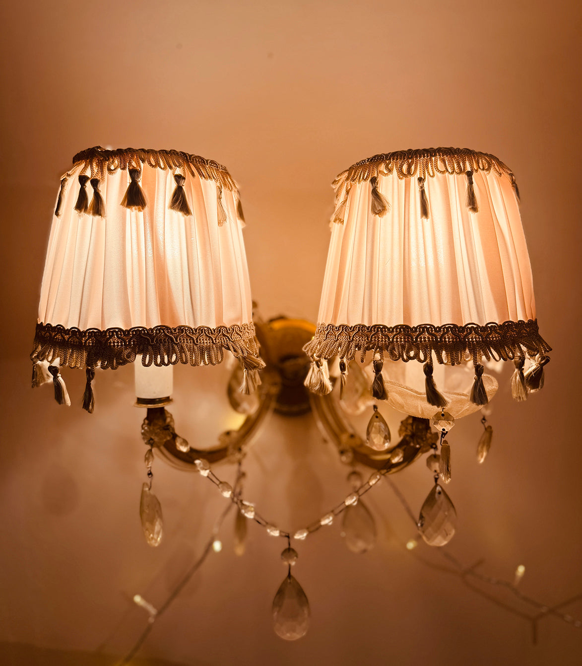 A double-arm wall sconce with two cream pleated fabric shades trimmed with dark tassel fringe, mounted on a warm brass-toned wall. Clear teardrop crystal drops hang from the arms, and the lamp is photographed against a warm beige wall.