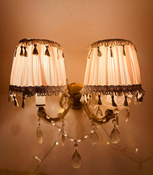 A double-arm wall sconce with two cream pleated fabric shades trimmed with dark tassel fringe, mounted on a warm brass-toned wall. Clear teardrop crystal drops hang from the arms, and the lamp is photographed against a warm beige wall.