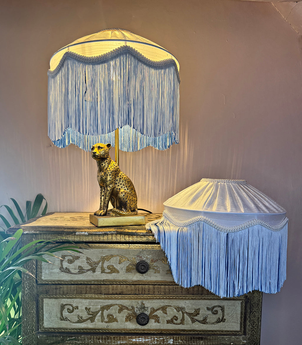 A decorative handmade white lampshade with blue and white fringe, displayed on an ornate vintage-style chest of drawers next to another matching fringed lampshade.